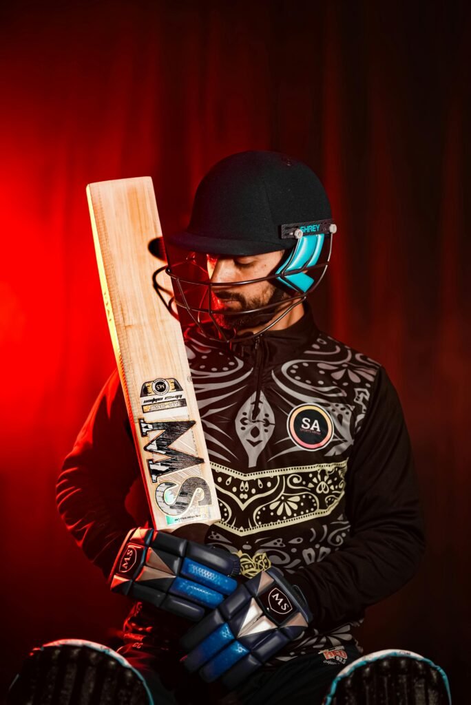 Portrait of a cricket player wearing gear, holding a bat under red lighting. Classic sports style.